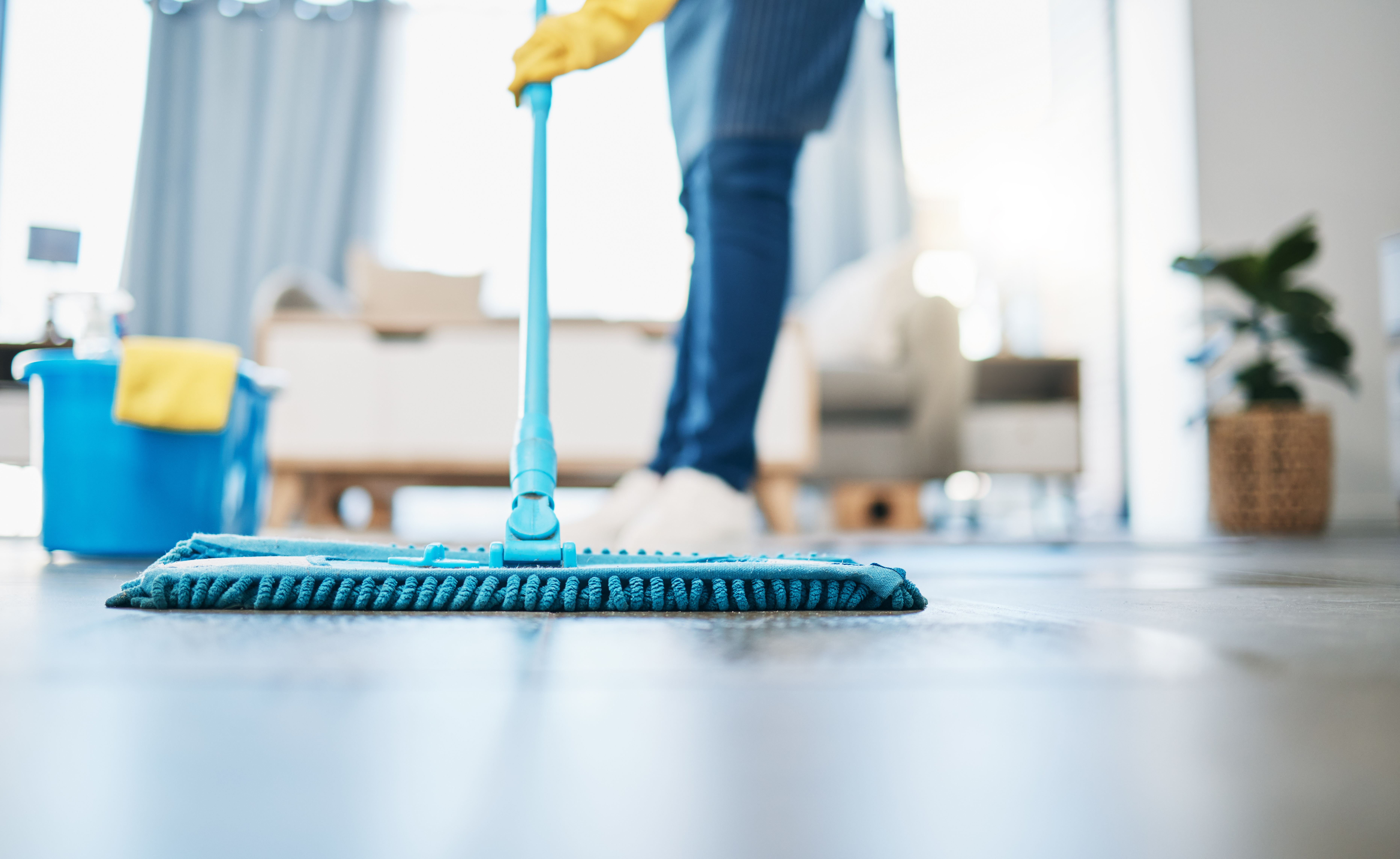Person mopping floor with blue mop and yellow gloves in bright living room