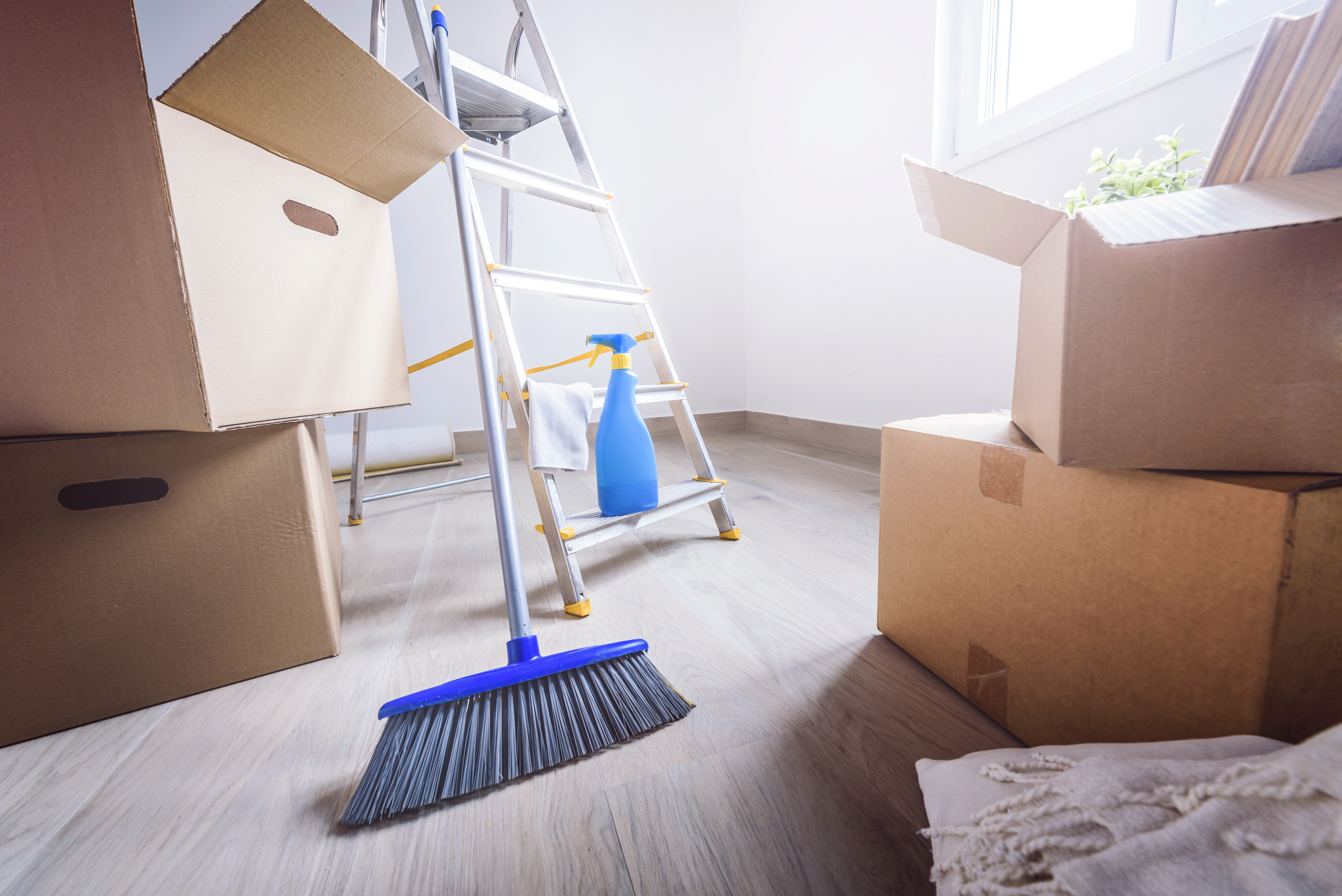 Moving day cleanup scene with cardboard boxes, ladder, broom, and spray bottle in empty room