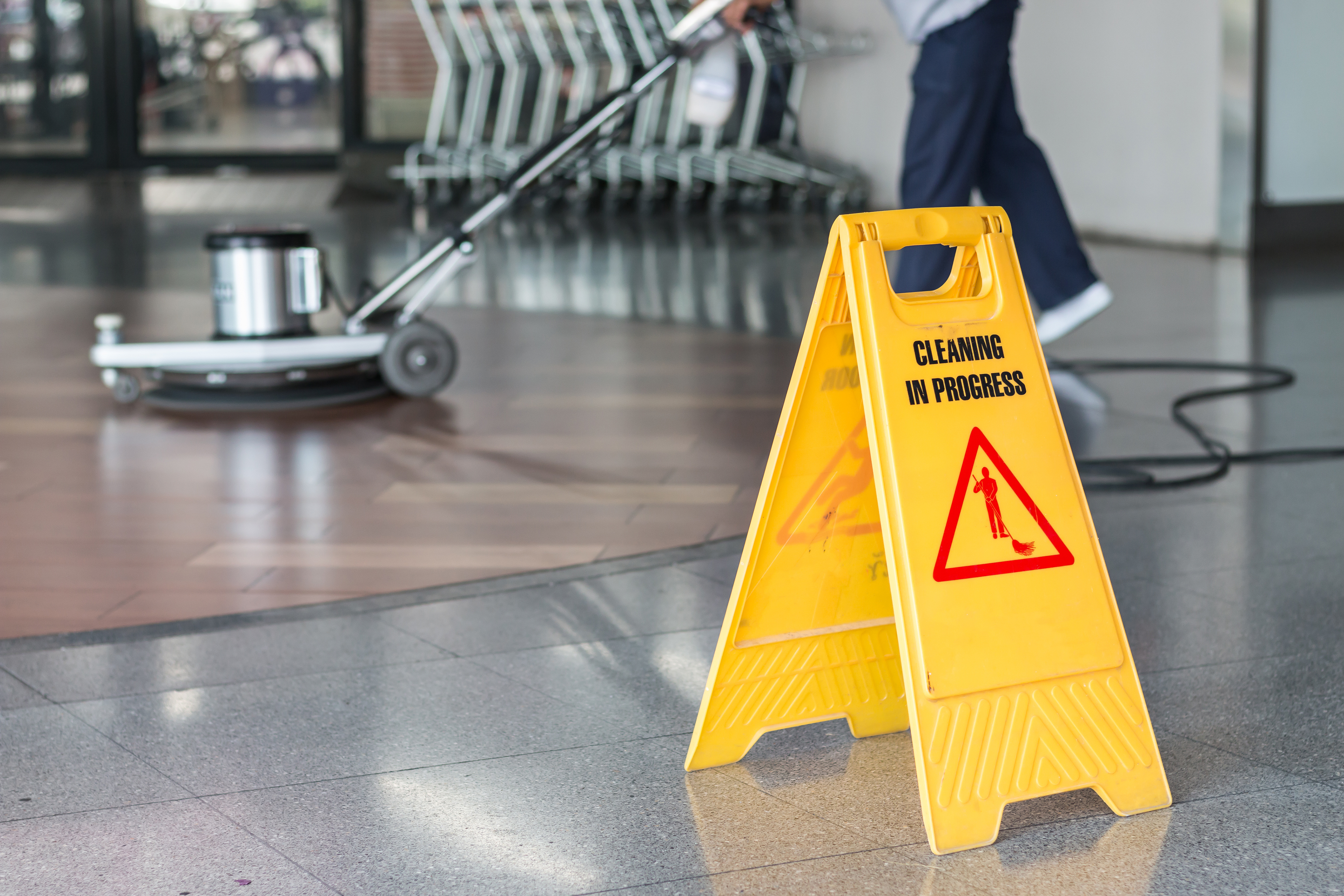 Yellow wet floor sign reading "Cleaning in Progress" with worker and equipment in background