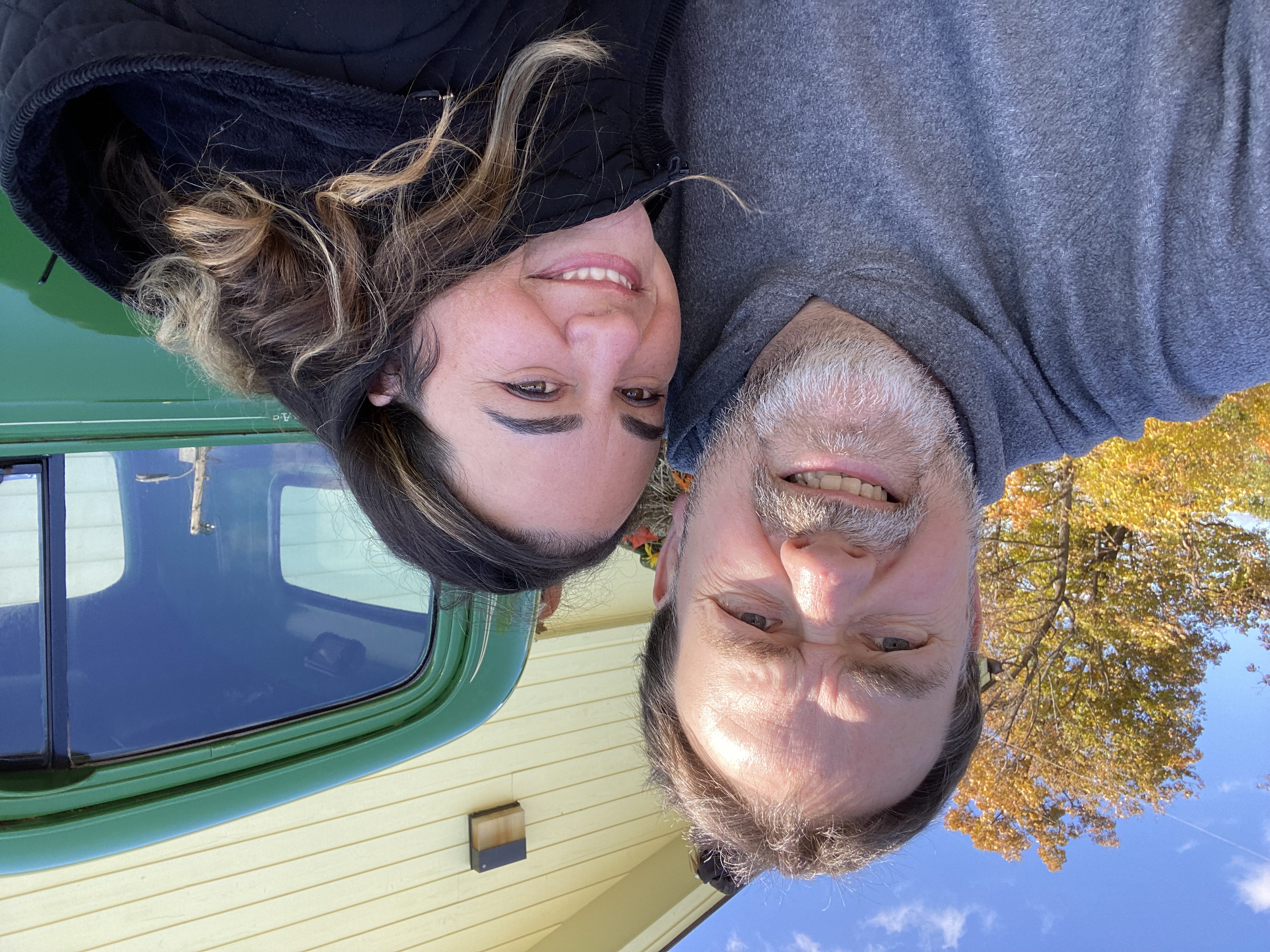 Two people smiling at camera in front of green RV and autumn trees